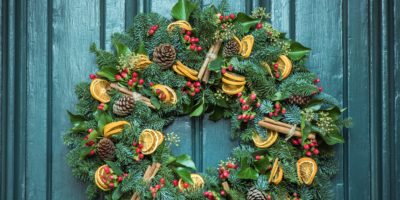 A christmas wreath with pine cones, dried slices of orange, holly and bundles of cinnamon sticks hanging below the brass knocker on a green front door.