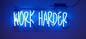 A pale blue neon sign that says, 'work harder'.