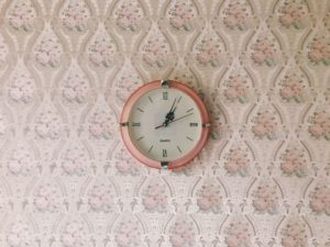 A pink clock on a pink floral patterned wall.