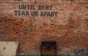 The words "until debt tear us apart" stencilled on a brick wall in black capital letters.