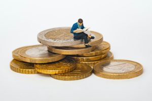 A stack of gold and silver coloured Euro coins with a small person wearing a blue jacket perched on the edge, reading a newspaper